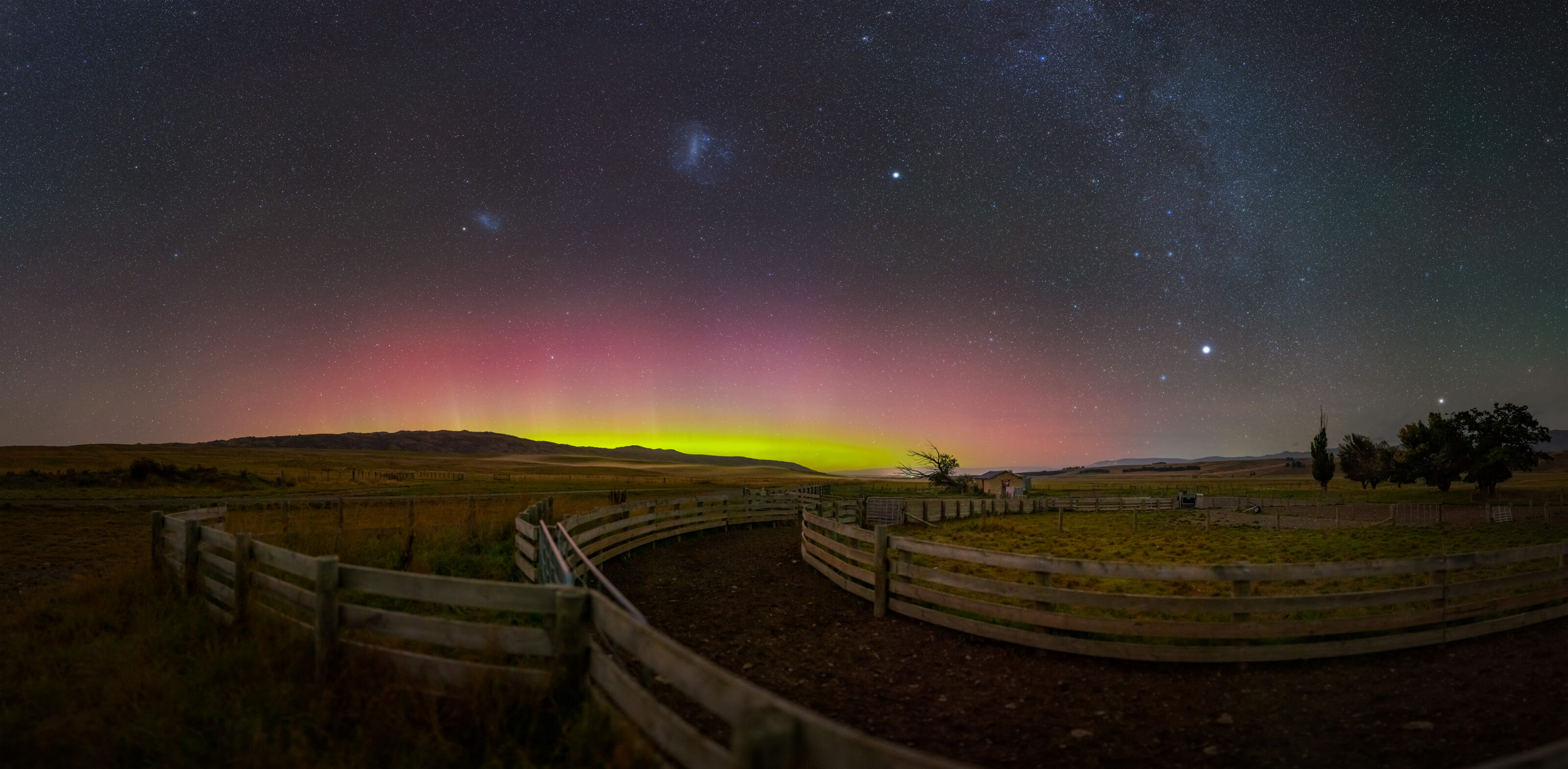 bright gree and pink Aurora Australis display below the starry sky with Large Megallanic Cloud, and above the foreground of Central Otago farm stockyards.