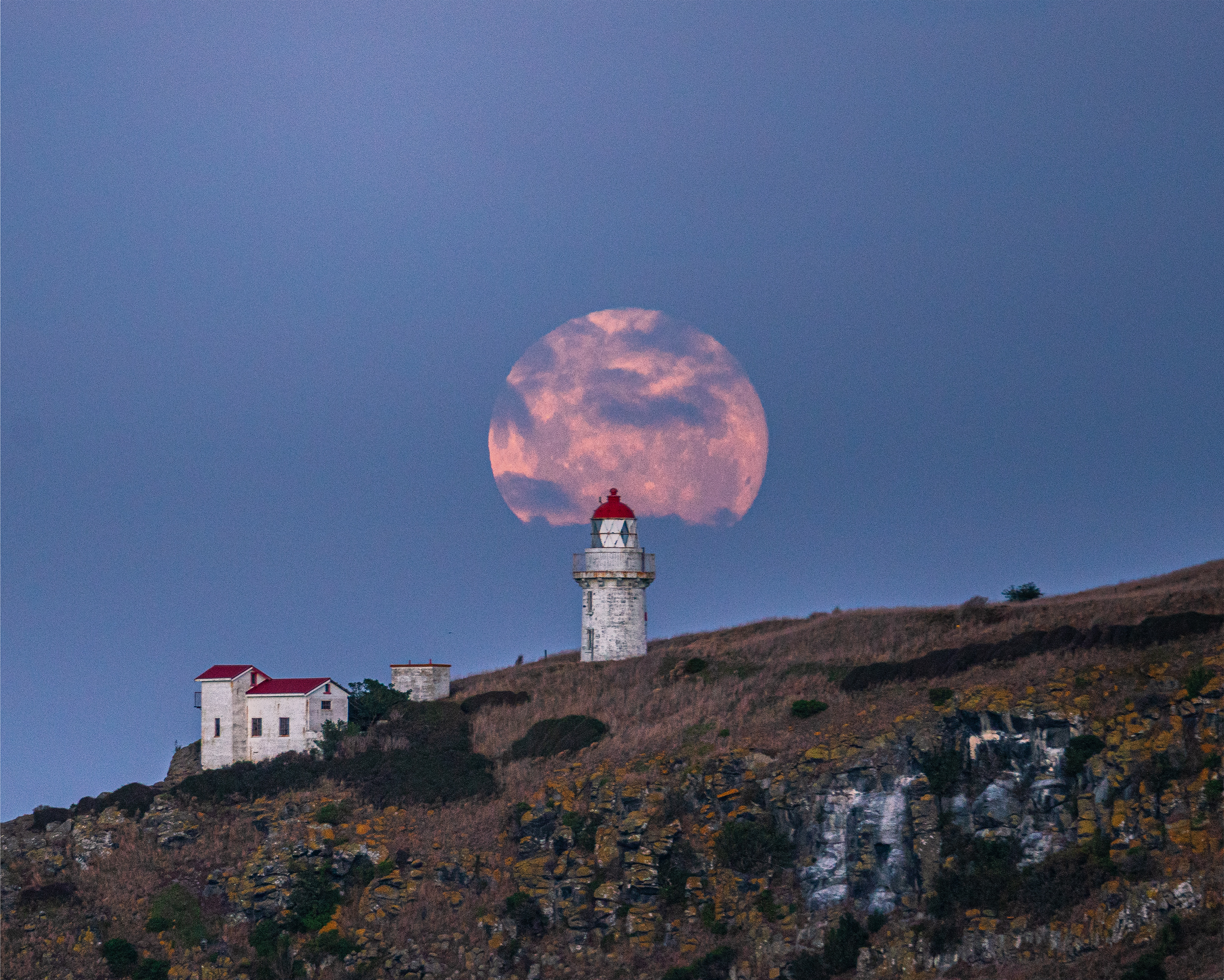 Telephoto lens shot zoomed in on lighthouse from Aramoana beach accross the Otago Harbour. The distance to shot gives effect of lighthouse dwarfed by full moon appearing twice as big behind it.