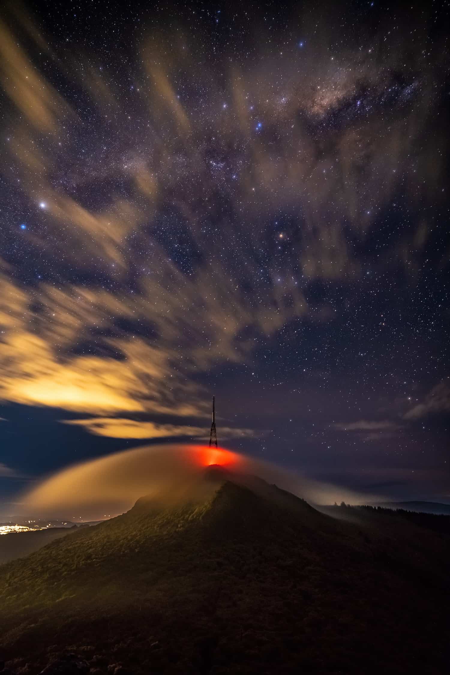 Red glow from the Mt Cargill communications tower shines through wind swept shield of cloud over the bush clad mountain. The lights of Dunedin city can be seen beyond and below the hill. Thi lights of the milky way above partially obscured by light cloud.