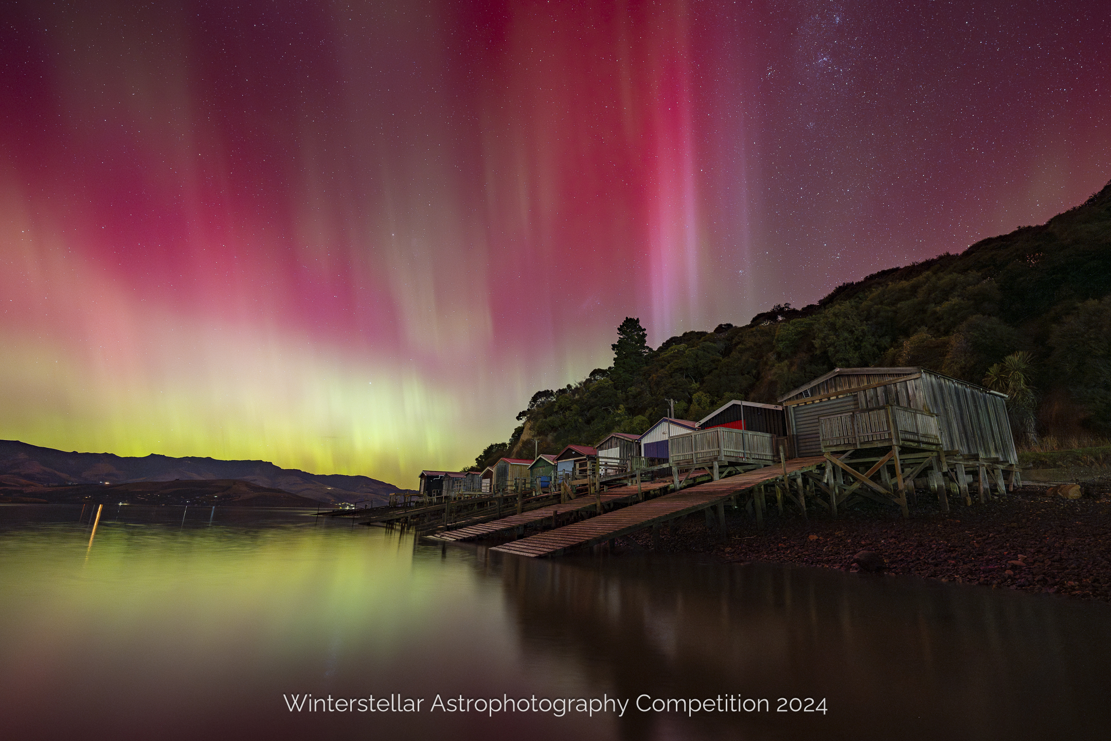 Stunning Aurora fills sky behind boat houses and water in foreground.