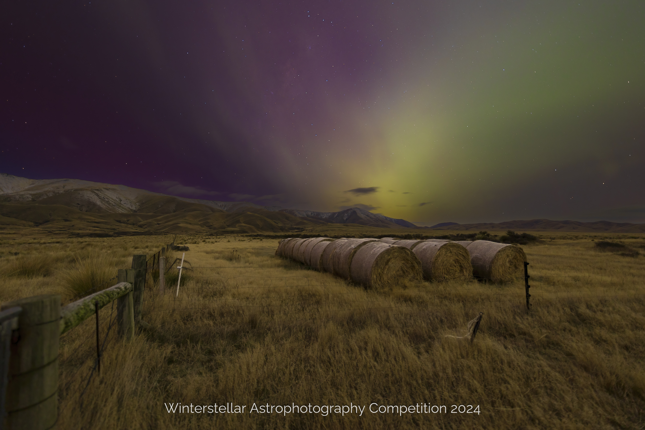 Green glow of an Aurora over distant hills and line of round hay bails.